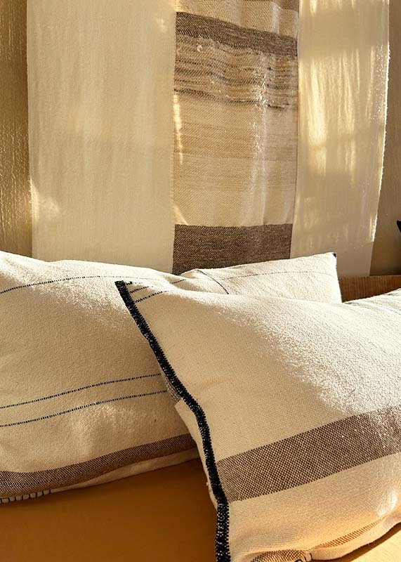 Two luxurious, handwoven natural cotton cushions sit against an original handwoven artwork in the afternoon light